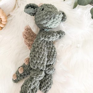 Peut inclure: Un dragon en crochet fait &agrave; la main, dans des tons de gris et de beige. Le dragon a de grandes oreilles, de petites ailes et des pieds arrondis. La peluche est pos&eacute;e sur une surface blanche et duveteuse.