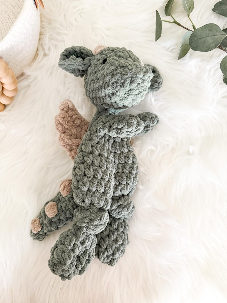 Peut inclure: Un dragon en crochet fait &agrave; la main, dans des tons de gris et de beige. Le dragon a de grandes oreilles, de petites ailes et des pieds arrondis. La peluche est pos&eacute;e sur une surface blanche et duveteuse.