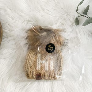 May include: A beige knitted beanie with a white pom pom and the word "Hi" knitted in white on the front. The beanie is packaged in a clear plastic bag with a sticker that says "Thank you".