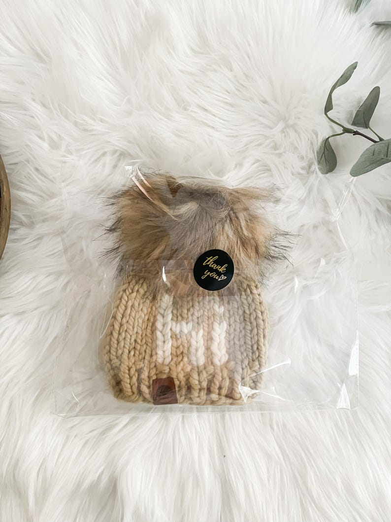 May include: A beige knitted beanie with a white pom pom and the word "Hi" knitted in white on the front. The beanie is packaged in a clear plastic bag with a sticker that says "Thank you".