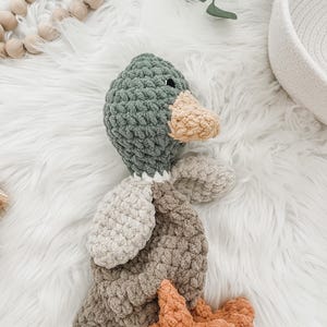 May include: A crocheted plush toy duck with a green head, beige body, and orange feet. The duck is lying on a white fluffy surface.