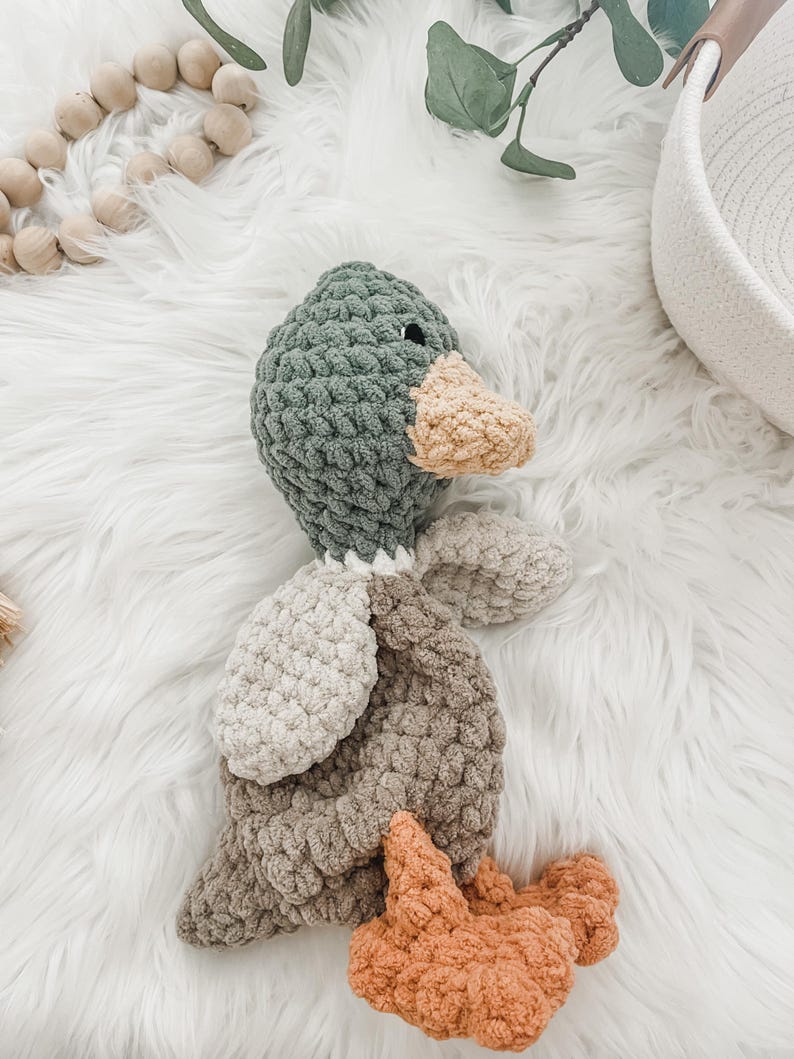 May include: A crocheted plush toy duck with a green head, beige body, and orange feet. The duck is lying on a white fluffy surface.