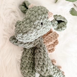Peut inclure: Un jouet dragon en peluche fait &agrave; la main, dans des tons vert sauge et beige. Le dragon pr&eacute;sente un design en tricot &eacute;pais et textur&eacute;, avec de petits yeux noirs et des accents beiges sur ses cornes et ses ailes. Le jouet est pos&eacute; sur une surface blanche et moelleuse.