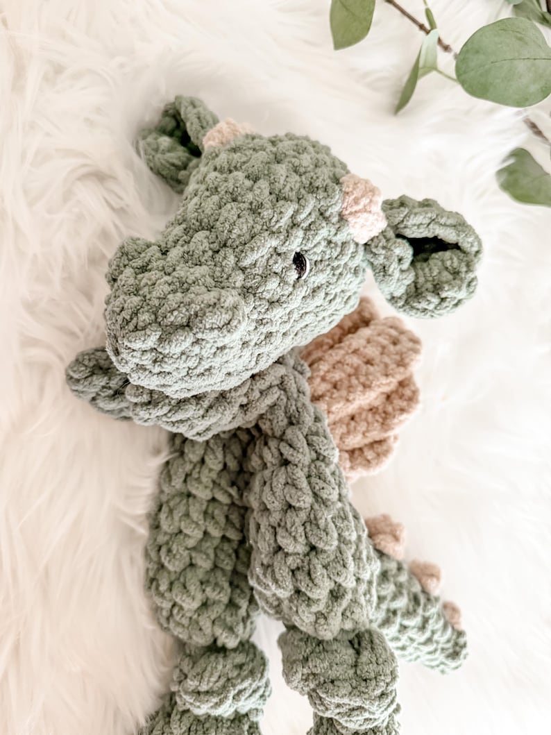 Peut inclure: Un jouet dragon en peluche fait &agrave; la main, dans des tons vert sauge et beige. Le dragon pr&eacute;sente un design en tricot &eacute;pais et textur&eacute;, avec de petits yeux noirs et des accents beiges sur ses cornes et ses ailes. Le jouet est pos&eacute; sur une surface blanche et moelleuse.
