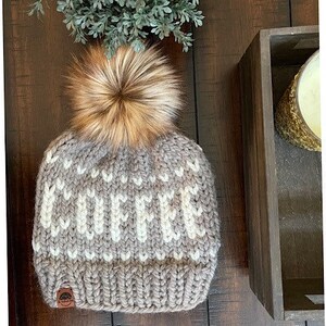 May include: A chunky knit beanie in shades of gray and cream, topped with a fluffy brown faux fur pom. The word "COFFEE" is knit into the hat. A small leather tag is sewn on the brim. A cozy winter accessory.