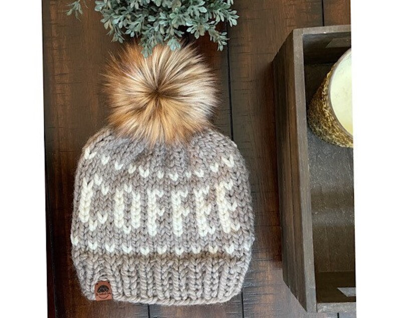 May include: A chunky knit beanie in shades of gray and cream, topped with a fluffy brown faux fur pom. The word "COFFEE" is knit into the hat. A small leather tag is sewn on the brim. A cozy winter accessory.