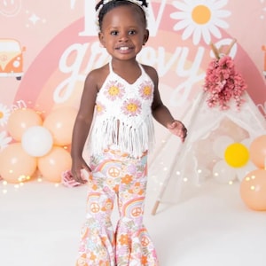 May include: A young girl wearing a white crocheted halter top with pink flowers and a white crocheted fringe. She is also wearing bell bottom pants with a rainbow, peace sign, and flower pattern. The background is a pink and peach ombre with a rainbow and a daisy.