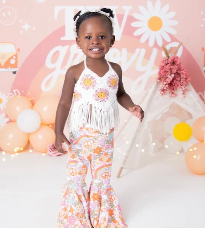 May include: A young girl wearing a white crocheted halter top with pink flowers and a white crocheted fringe. She is also wearing bell bottom pants with a rainbow, peace sign, and flower pattern. The background is a pink and peach ombre with a rainbow and a daisy.