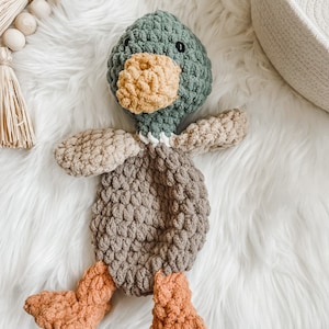 May include: A crocheted duck plush toy with a green head, brown body, and orange feet. The toy is lying on a white fluffy surface.