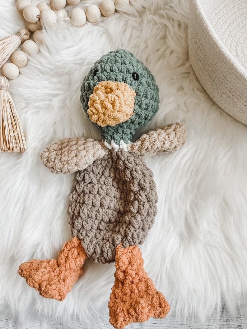 May include: A crocheted duck plush toy with a green head, brown body, and orange feet. The toy is lying on a white fluffy surface.