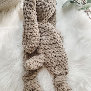 May include: A brown crocheted bunny plush toy lying on a white fluffy surface.