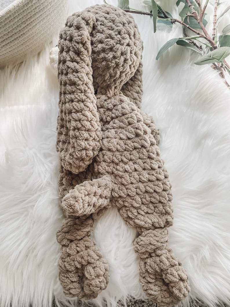 May include: A brown crocheted bunny plush toy lying on a white fluffy surface.