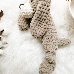 May include: A crocheted plush dog toy in brown and white with a long body and floppy ears. The toy is lying on a white fluffy surface.