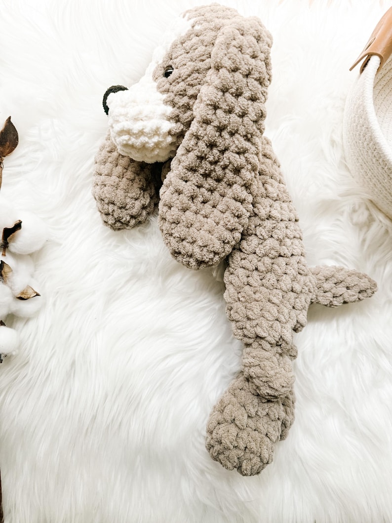 May include: A crocheted plush dog toy in brown and white with a long body and floppy ears. The toy is lying on a white fluffy surface.