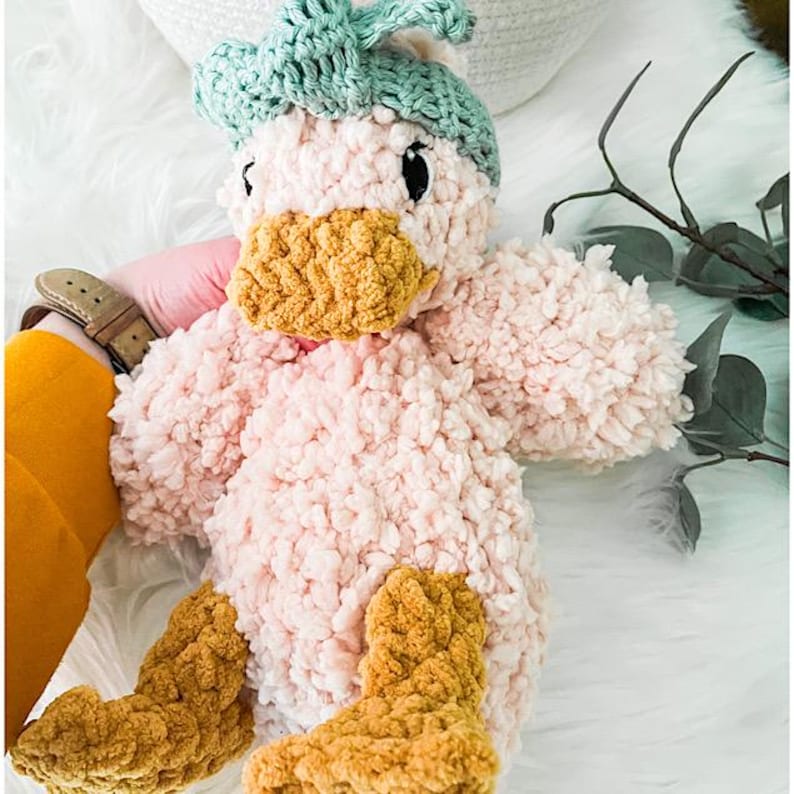 May include: A pink plush duck toy with a crocheted green hat and brown feet. The duck has a fluffy texture and a friendly expression.