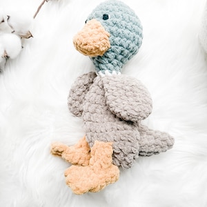 May include: A handmade crochet duck toy in shades of blue, gray, and tan. The duck has a blue head, a tan beak and feet, and gray body and wings. The toy is lying on a white, fluffy surface.