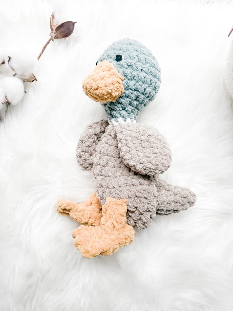 May include: A handmade crochet duck toy in shades of blue, gray, and tan. The duck has a blue head, a tan beak and feet, and gray body and wings. The toy is lying on a white, fluffy surface.