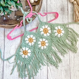 May include: A green crochet halter top with white and yellow daisy flowers and fringe trim.