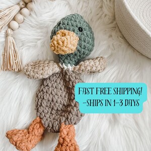 May include: A handmade crochet duck plushie in shades of green, tan, brown, and orange. The duck has a textured body, wings, and feet. The duck's head is green with a yellow beak and black eyes. The plushie is on a white fluffy surface.