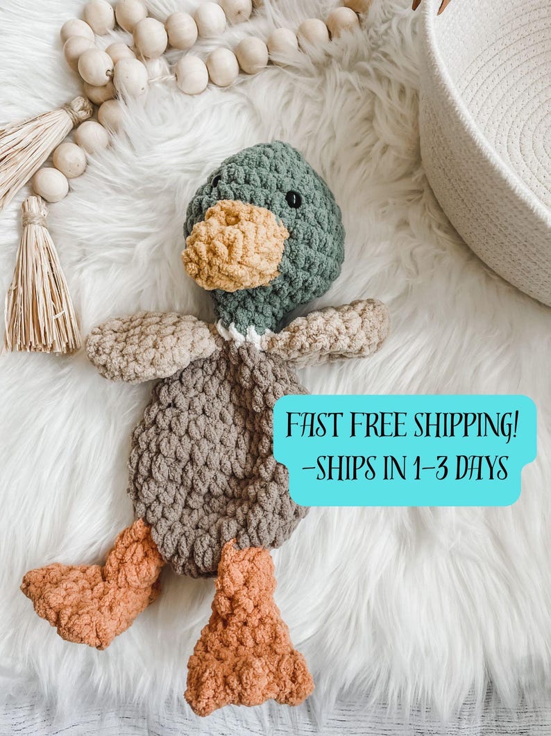 May include: A handmade crochet duck plushie in shades of green, tan, brown, and orange. The duck has a textured body, wings, and feet. The duck's head is green with a yellow beak and black eyes. The plushie is on a white fluffy surface.