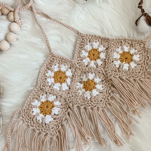 May include: A crocheted halter top with a daisy pattern and fringe. The top is made in a beige color with white and yellow accents.