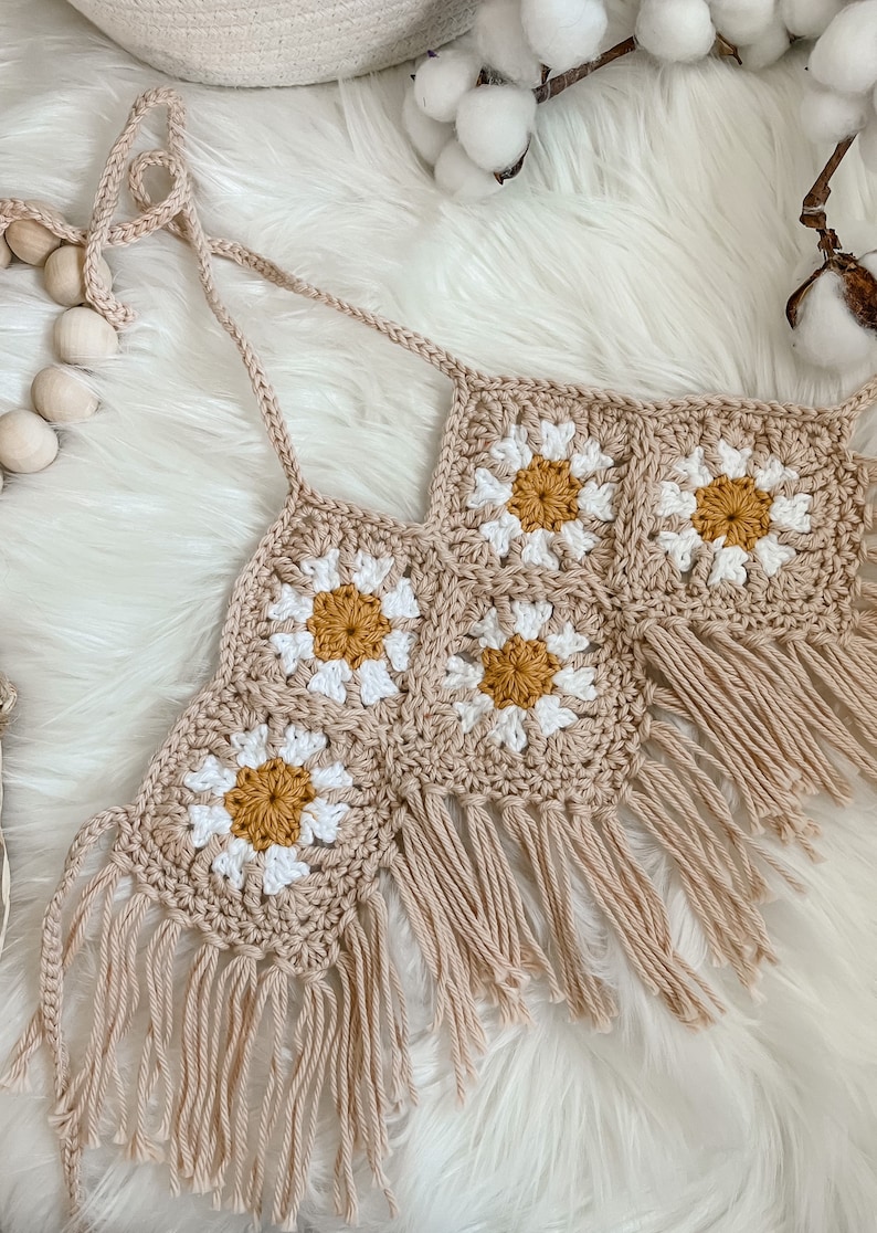 May include: A crocheted halter top with a daisy pattern and fringe. The top is made in a beige color with white and yellow accents.