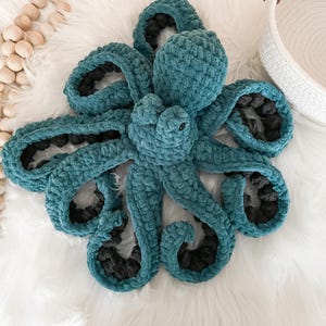 Pode incluir: Um brinquedo de pel&uacute;cia de polvo de croch&ecirc; azul-turquesa com detalhes pretos. O polvo tem oito tent&aacute;culos e est&aacute; deitado em uma superf&iacute;cie branca e fofa.