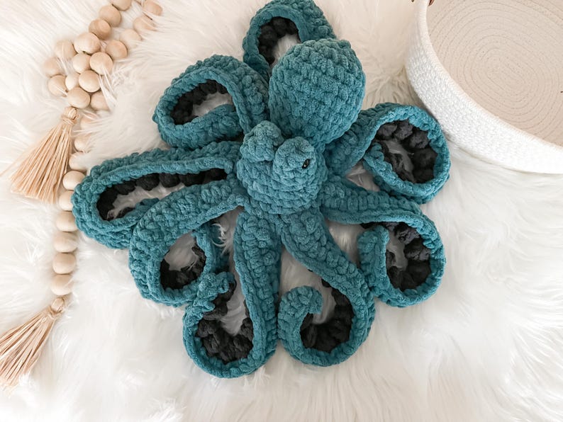 Pode incluir: Um brinquedo de pel&uacute;cia de polvo de croch&ecirc; azul-turquesa com detalhes pretos. O polvo tem oito tent&aacute;culos e est&aacute; deitado em uma superf&iacute;cie branca e fofa.