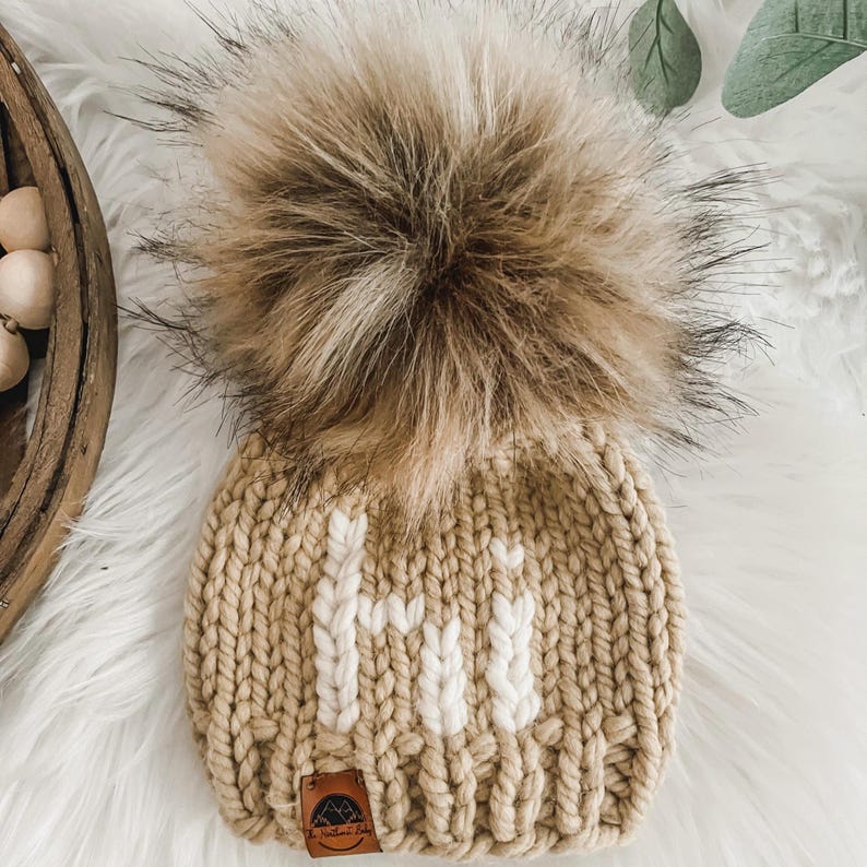 May include: A beige knitted hat with a white "Hi" embroidered on the front. The hat has a large brown faux fur pom pom on top.