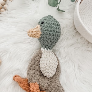 May include: A crocheted duck toy with a green head, brown body, and orange feet. The duck is lying on a white fluffy surface.