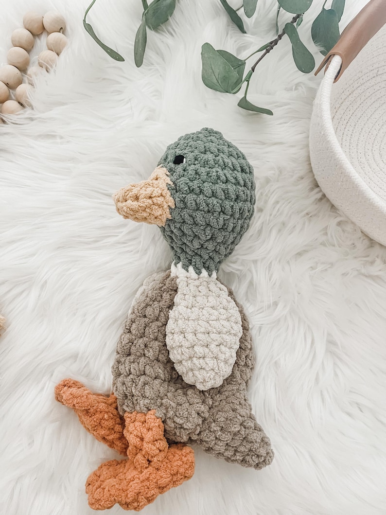May include: A crocheted duck toy with a green head, brown body, and orange feet. The duck is lying on a white fluffy surface.