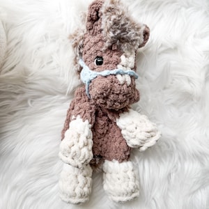 May include: A handmade crochet horse toy in shades of brown and cream. The horse has a fluffy mane and tail, a blue bridle, and black eyes. The toy is made of soft yarn and is designed to be a cuddly companion.
