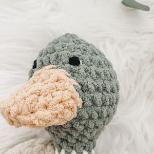 May include: A crocheted plush toy shaped like a duck with a gray head and a beige beak. The duck is lying on a white fluffy surface.