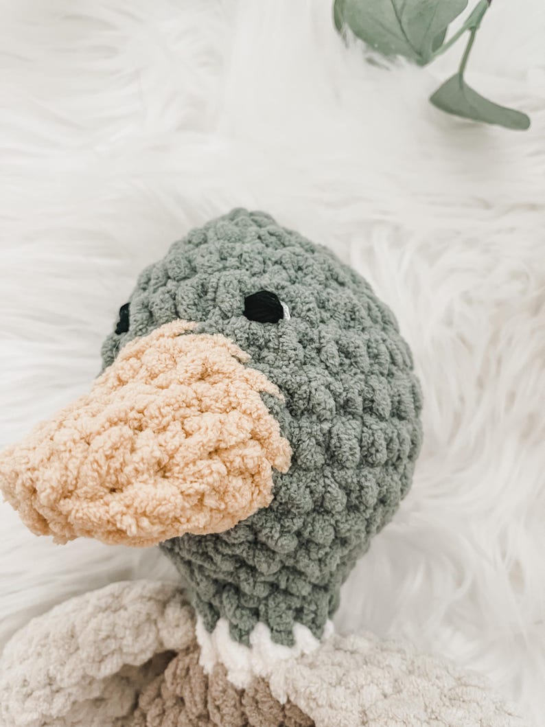 May include: A crocheted plush toy shaped like a duck with a gray head and a beige beak. The duck is lying on a white fluffy surface.