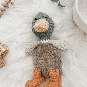 May include: A crocheted plush toy duck with a green head, brown body, and orange feet. The duck is sitting on a white fluffy surface.