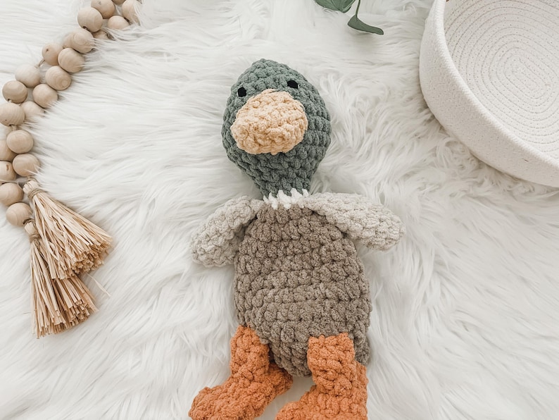 May include: A crocheted plush toy duck with a green head, brown body, and orange feet. The duck is sitting on a white fluffy surface.