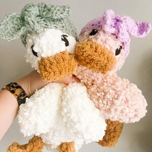 May include: Two crocheted plush duck toys. One duck is white with a sage green crocheted hat. The other duck is pink with a lavender crocheted hat. Both ducks have brown beaks and feet.