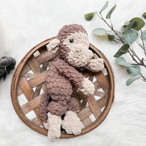 May include: A handmade crochet monkey in shades of brown and tan sits in a round, woven basket. The plush toy has a light tan face, hands, and feet, with a darker brown body. The basket is made of light brown wood.