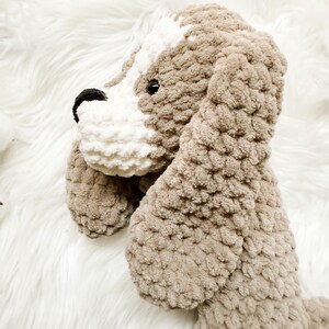 May include: A crocheted plush dog toy with brown and white fur. The dog is sitting up with its head tilted to the side. The toy is made with a soft, fluffy yarn.