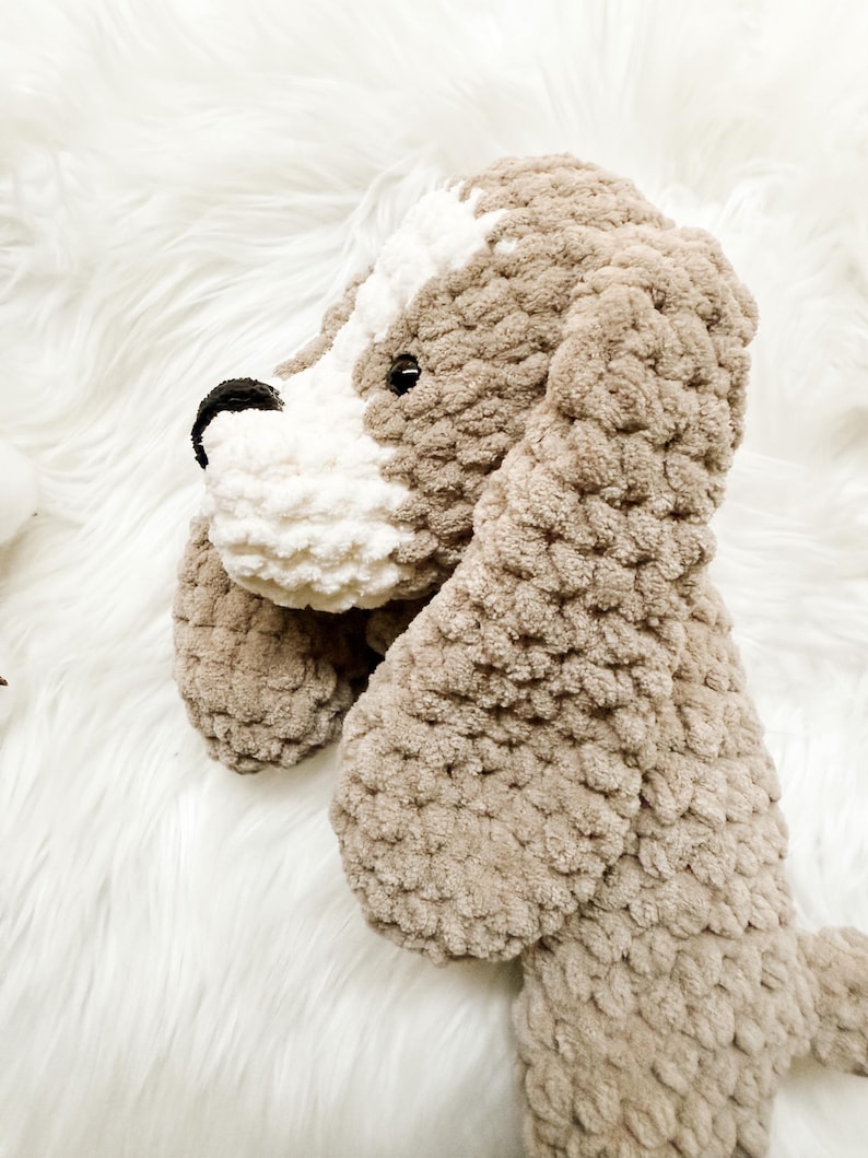 May include: A crocheted plush dog toy with brown and white fur. The dog is sitting up with its head tilted to the side. The toy is made with a soft, fluffy yarn.