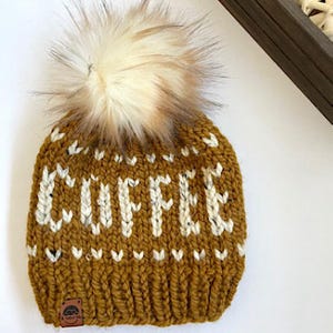 May include: Mustard yellow knit beanie with the word "COFFEE" in white block letters. The hat features a large, fluffy, cream-colored pom-pom on top. A small leather tag is sewn on the brim.