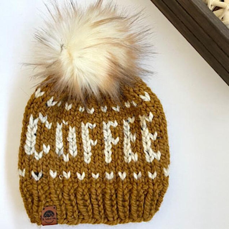 May include: Mustard yellow knit beanie with the word "COFFEE" in white block letters. The hat features a large, fluffy, cream-colored pom-pom on top. A small leather tag is sewn on the brim.