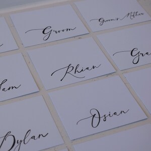 May include: Square white place cards with elegant black calligraphy. Names like "Bride," "Groom," and individual names are written in a flowing script. Ideal for weddings or special events.