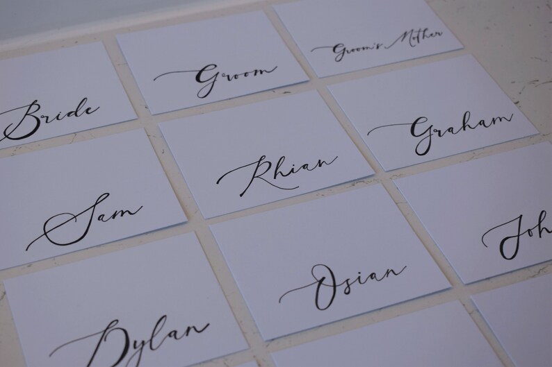 May include: Square white place cards with elegant black calligraphy. Names like "Bride," "Groom," and individual names are written in a flowing script. Ideal for weddings or special events.
