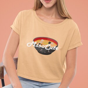 May include: A woman wearing a light beige cropped t-shirt with a cartoon miso soup bowl and the text "Miso Cute" printed on it. She is sitting on a wooden chair with her legs crossed.