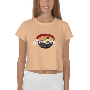 May include: A peach colored cropped t-shirt with short sleeves featuring a cartoon illustration of a bowl of miso soup with the text "MisoCute" in white.