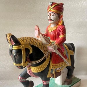 May include: A hand-painted wooden figurine depicting a person riding a black horse. The rider wears a red and gold outfit and a turban. The horse has gold and pink accents. The figurine is mounted on a green and white base.