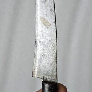 Puede incluir: Un cuchillo antiguo con una hoja curvada y desgastada y un mango de madera oscura. La hoja muestra signos de uso y edad, con un acabado ligeramente opaco. El mango es de color marrón oscuro y parece ser de madera.