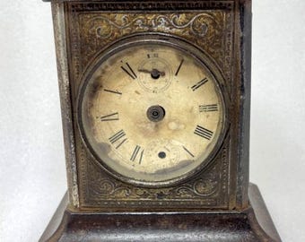 Könnte beinhalten: Antike Metalluhr mit rechteckigem Sockel und aufwendigen Verzierungen. Das Zifferblatt zeigt römische Ziffern und zwei kleinere Zifferblätter. Ein Griff ist oben angebracht. Die Uhr hat ein verwittertes, gealtertes Aussehen.