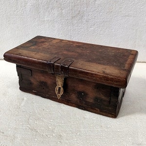 Puede incluir: Pequeña caja rectangular de madera oscura con tapa con bisagras. La caja es de madera marrón oscura con vetas visibles y se asegura con un cierre metálico. La caja parece ser vintage o antigua.